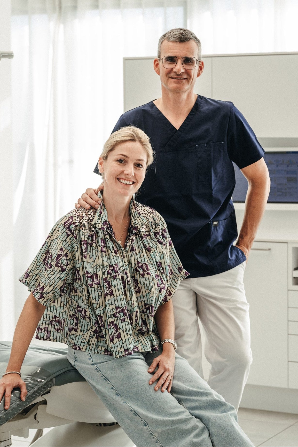 Two people in a dental office setting, with one person sitting on a chair and the other standing behind.